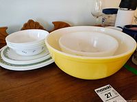 Photo showing two large Pyrex bowls, one yellow and one white nested inside, with white floral bowls and Corelle plates stacked to the side.