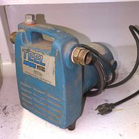 Side view of blue Teel water system pump showing handle, brass pipe connections and attached power cord with plug.