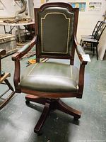 Front angle view of wooden rolling executive chair with green leather upholstery and gold decorative border, showing overall condition and armrests