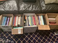 Five cardboard boxes filled with books arranged on floor by a sofa