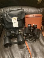 Lot showing two pairs of binoculars with respective carrying cases on couch, one leather brown case and one black nylon case.