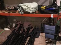 Two folded hunting-style camping chairs with camouflage fabric and black legs, along with some folded and rolled camping gear, bags, and camping pads on a wooden pallet in a storage area.