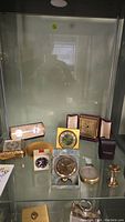 Glass shelf displaying nine small timekeeping devices including folding clocks, gauge and hourglass