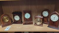 Six different wooden mantel clocks arranged on a wood shelf, view of faces and cases