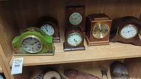 View of six assorted mantel clocks on wood shelves. Clocks vary in shape, size and wood tone with different dial styles.