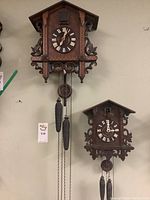Photo showing two traditional wall-hanging cuckoo clocks mounted on a wall with pinecone weights and carved leaf details.