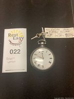 Overall view of pocket watch and winding key next to Rest Easy tag