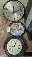 Three wall clocks stacked: modern silver-faced clock, Elvis Presley themed clock, traditional clock with Roman numerals.