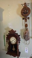 Two clocks displayed: a carved wood-cased pendulum clock on the left and a wooden foliot-style clock kit on the right.