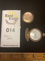 Two views of the pocket watch showing closed and open covers alongside a lot identifier card and ruler.