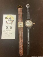 Two wrist watches including a vintage Mickey Mouse watch with square face, and an antique round-face watch on black leather strap alongside a lot tag.