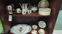 Photo showing a wood cabinet shelf with a white and black trimmed tea pot, matching tea cups and saucers, clock face small plates, and a porcelain sugar bowl, along with several decorative plates and a large oval serving platter.