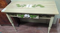 Side and partial top view of table highlighting the floral painting on drawer and natural wood finish.