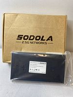 Closed cardboard box labeled 'SODOLA 2.5G NETWORKS' and the Ethernet switch inside a protective plastic bag with a white information label on the back.