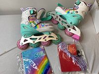 Pair of children's inline roller skates showing side and top views on a flat surface including accessories: purple laces, red pouch with tool, and rainbow-branded plastic bag.