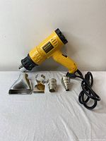 Yellow electric heat tool with four different metal nozzles, placed on white surface against light wall.