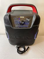 Front view of Ion Pathfinder 360 Bluetooth speaker with illuminated control panel, red handle, and included power cord placed in front.