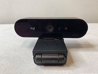 Front view of Logi webcam showing camera lens, built-in microphone, and LED light on glossy black surface, mounted on folded clip.