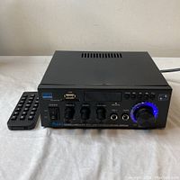 Front view of AK-45 amplifier with remote control showing power switch, treble, bass, mid/vol knobs, microphone inputs, USB port, LED display, mode buttons, and illuminated volume knob.