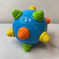 Photo of colorful blue plastic spherical shake ball toy with yellow, orange, and green textured knobs. Front view showing detailed embossed shapes on the ball and textures on each knob.