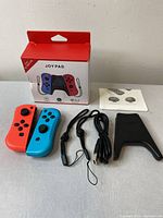 Joypad set contents displayed on table including red and blue joypads, wrist straps, charging cable, mounting accessory, and user manual with packaging box.