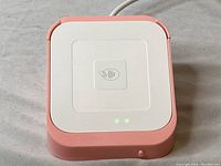 Front view of Square contactless payment reader device with pink base, white top, contactless payment symbol and two green LED lights active.