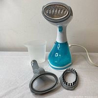 Front view of handheld garment steamer showing steamer head, measuring cup, and two attachments laid out on white surface.