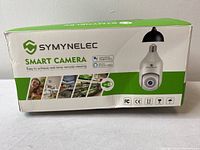 Front of the product box showing Symynelec Smart Camera branding, images illustrating use cases, Wi-Fi capability, and compatibility with Alexa and Google Assistant.
