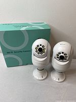 Front view of two white Wi-Fi security cameras with LED lights arranged around the lens and the original greenish packaging box behind them.