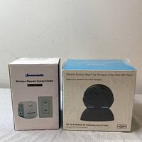 Box of Jewenwils Wireless Remote Control Outlet and boxed Mission Battery Base for Amazon Echo Dot (4th Gen) wrapped in plastic, shown side by side on a flat surface.