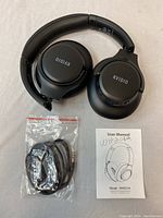 Photo showing closed foldable black KYDIO WH201A wireless headphones, user manual, and sealed cables pack