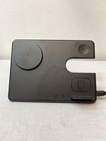 Top view of black rectangular 3-in-1 magnetic charging stand showing circular magnetic charging pad, rectangular area for smartwatch, and small rectangular dock for earbuds with cable attached.