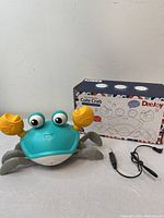 Photo of the dancing crab toy with teal body, large eyes, yellow claws, black power adapter, and original box.