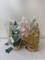 Five decorative Christmas trees in various colors and styles wrapped in plastic with tags, standing on wooden bases.