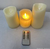 Photo showing three ivory-colored flameless candles of varying heights with one candle lit and a remote control in front.