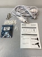 Photo of white outdoor extension cords with bulb sockets, metal S-hooks, white wire clips, plastic drywall anchors, and printed installation guide on gray surface.