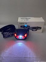 Front view of headlamp turned on with white and red LEDs illuminated, black and gray strap visible, original box in background