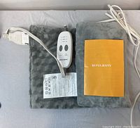 Grey massaging weighted heating pad folded with wired controller and yellow user manual on top.