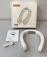 Fully unboxed neck fan kit showing the white ergonomic neck fan, USB charging cable, instruction manual, and retail box with product image.