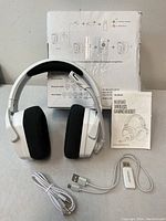 White Nubwo wireless gaming headset with microphone, USB dongle, USB charging cable, 3.5mm audio cable, and instruction manual displayed on a white surface with the headset box in background.