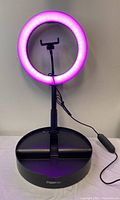 Front view of the illuminated ring light in pink color, mounted on black stand with phone holder. Power cord and inline control visible.