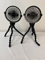 Front view of two black portable fans with circular grills on flexible tripod stands against a white background.