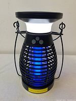 Front view of black caged solar powered bug zapper lantern showing white top light and blue UV insect attracting light inside cage.