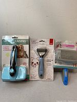 Three new pet grooming brushes in original packaging: a self-cleaning slicker brush, a small dog rake, and a fine-tooth comb, displayed side by side.
