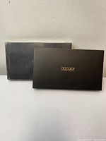 Two black rectangular boxes with gold Apodar logo on the front, one box in front and the other behind against a plain light wall.