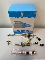 Box and items laid out: cat litter scoop box and three collars including plaid fabric collar with bell and bow tie, white collar with bell, pink collar in package.