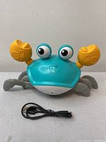 Front view of the electronic dancing crab toy showing turquoise body, yellow claw pincers, and black USB charging cable in front.