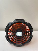 Front view of the small round fan showing the orange blades, black protective grille, and bright LED light in the center.