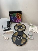 Full kit laid out showing box, two reels of LED strip lights on spools, power adapter, white controller, remote control, mounting clips, and user manual.