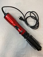 Full view of the red hairdryer brush with black plastic vented bristles and attached power cord, showing control buttons on the handle.